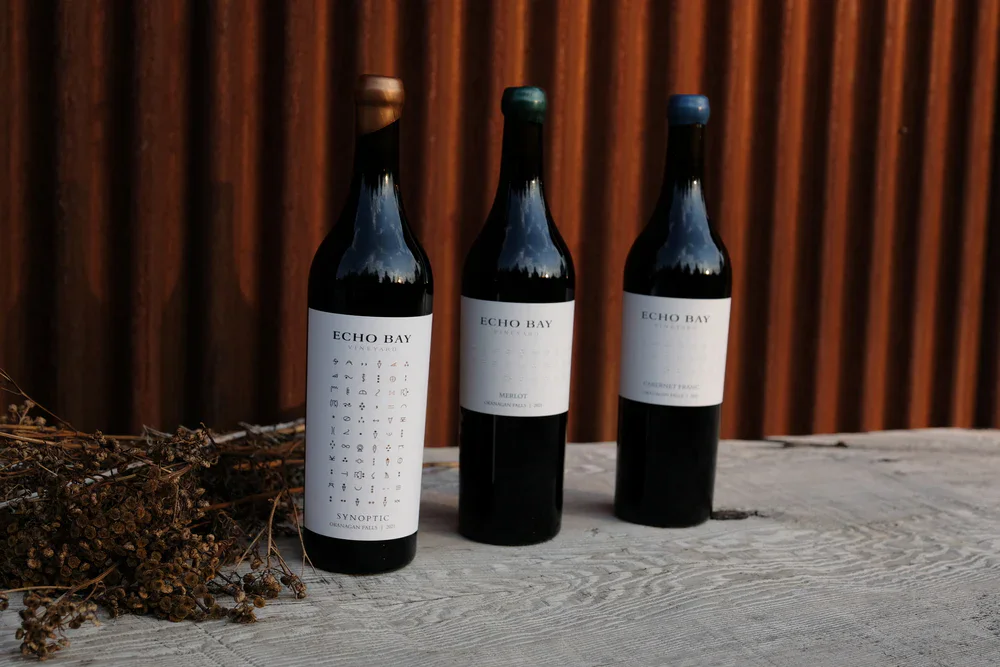 Three Echo Bay Vineyard wine bottles displayed on a rustic wooden surface with a wooden fence background.