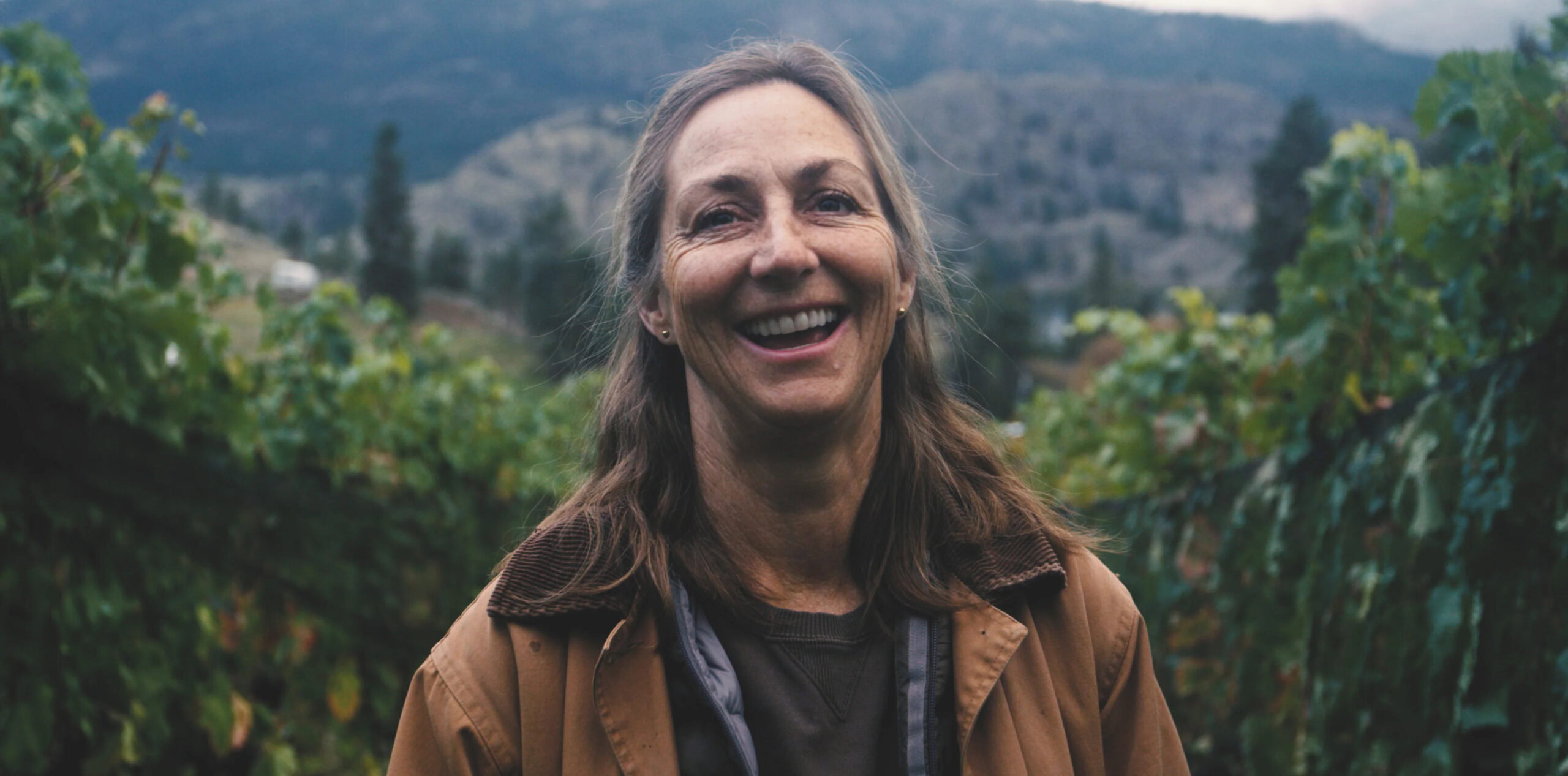 Vineyard landscape with a smiling woman in the foreground, lush grapevines, and scenic mountains in the background.