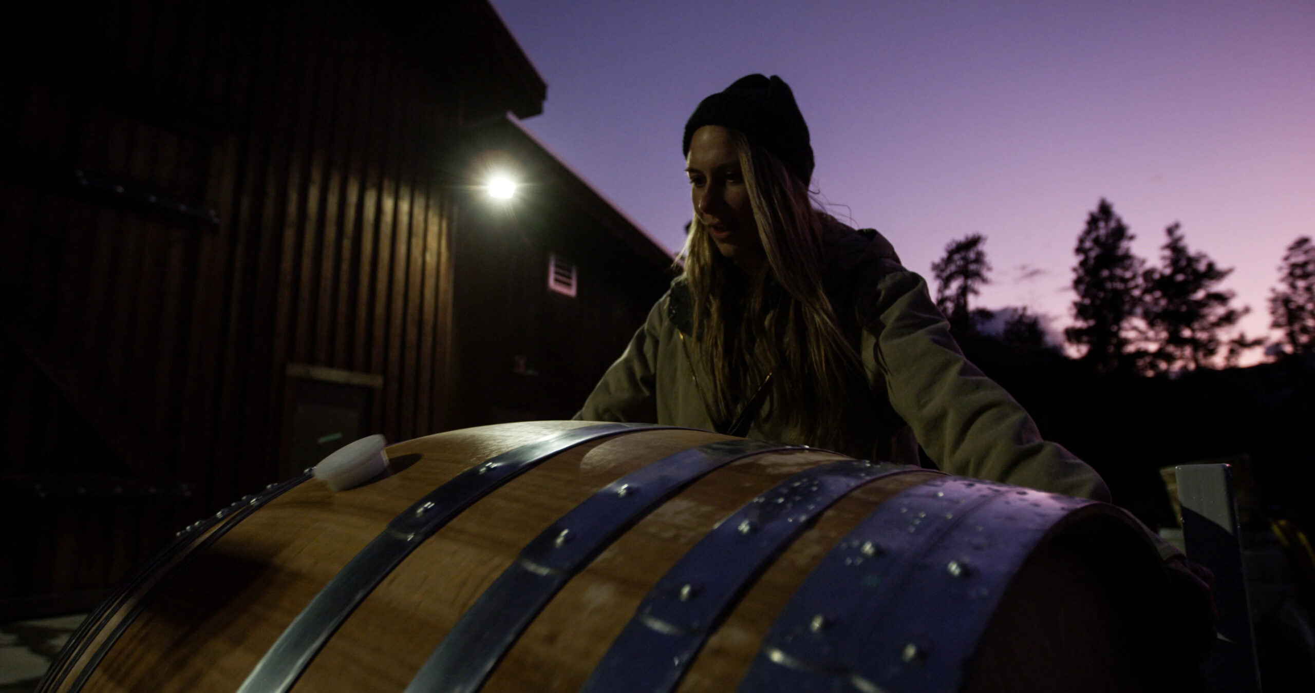 Kelsey Rufiange inspecting a wine barrel at Echo Bay Vineyard during sunrise, with a barn and trees in the background.